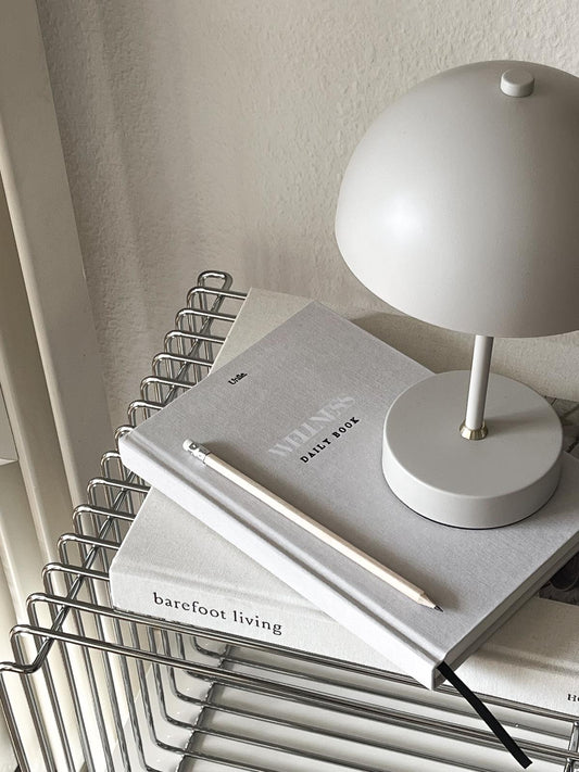Wellness book aesthetics Unile Self care journaling bien-être et santé posé de côté sur le livre Barefoot Living avec une lampe, un crayon beige et sur une étagère métallique.