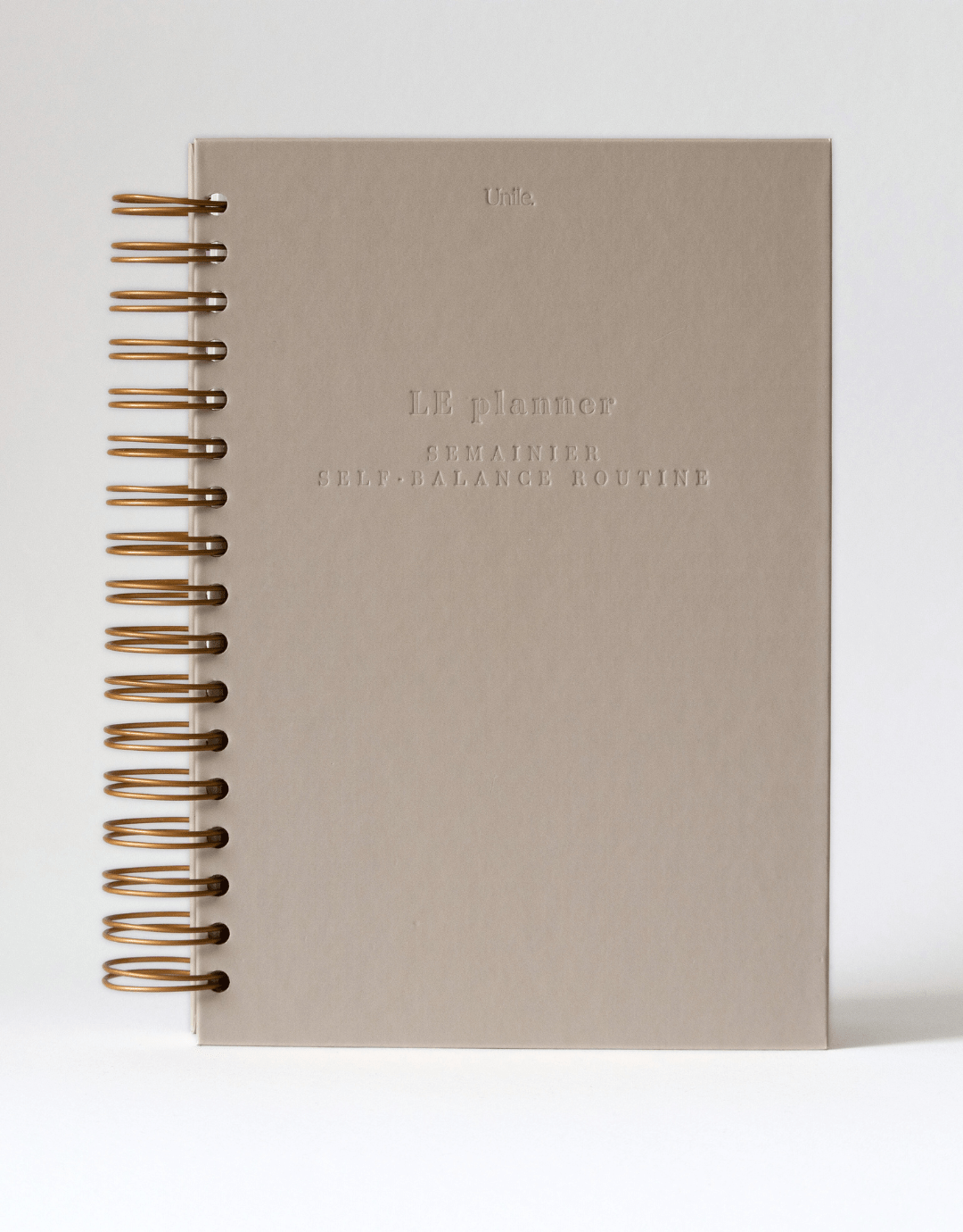 LE planner beige Unile Self-care planner avec grandes spirales dorées et lettres gravées.