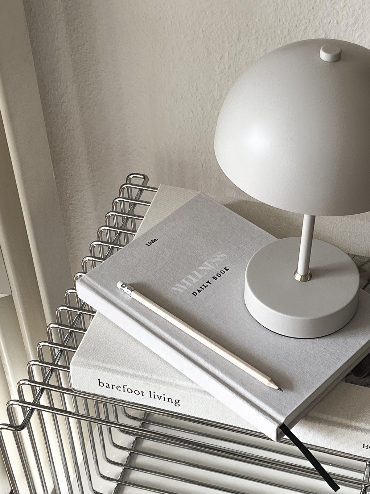 Wellness book aesthetics Unile Self care journaling bien-être et santé posé de côté sur le livre Barefoot Living avec une lampe, un crayon beige et sur une étagère métallique. 