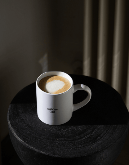 LA tasse - unile.selfcare tasse blanche lettrage noir Self-care Club posée sur un tabouret noir et jeux d'ombres en fond